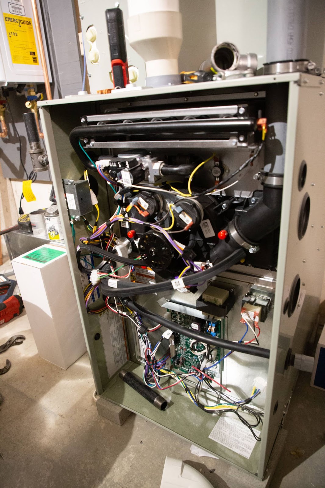 An open furnace unit with visible internal components and wires, situated in a basement or utility room. Tools and other equipment are partially visible in the background.