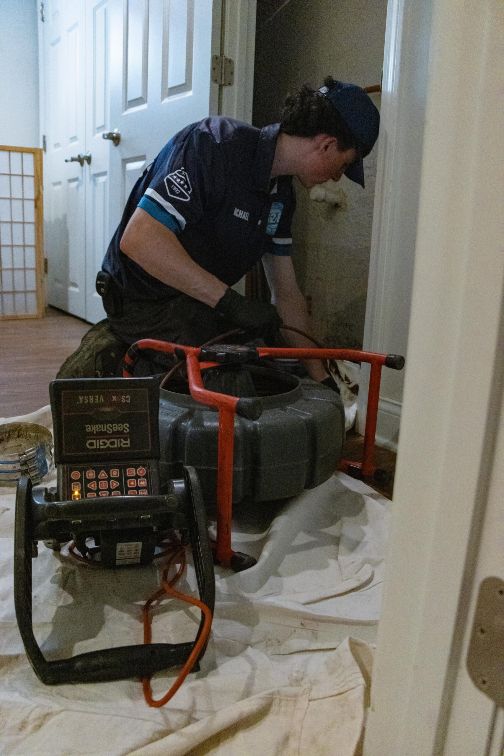 A technician in uniform uses plumbing inspection equipment on a pipe inside a residential home, with tools and a camera monitor on a protective sheet.