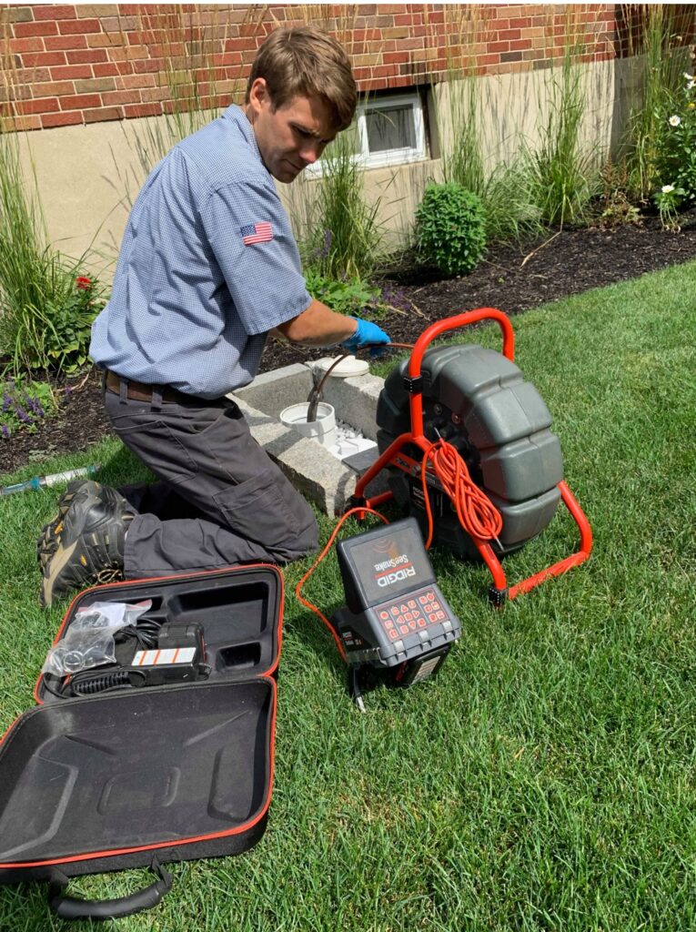 A technician kneels on grass, operating a pipe inspection camera system near a concrete access point outside a brick building.