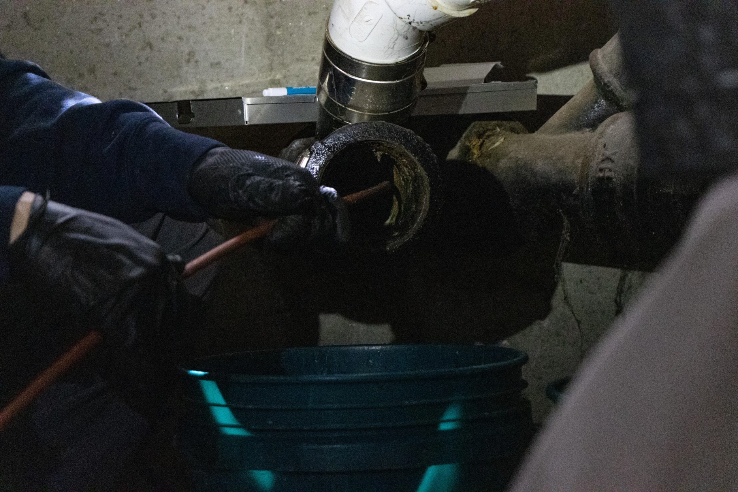 In a dimly lit space, a South Boston plumber in gloves diligently cleans a pipe with precision tools over a green bucket.