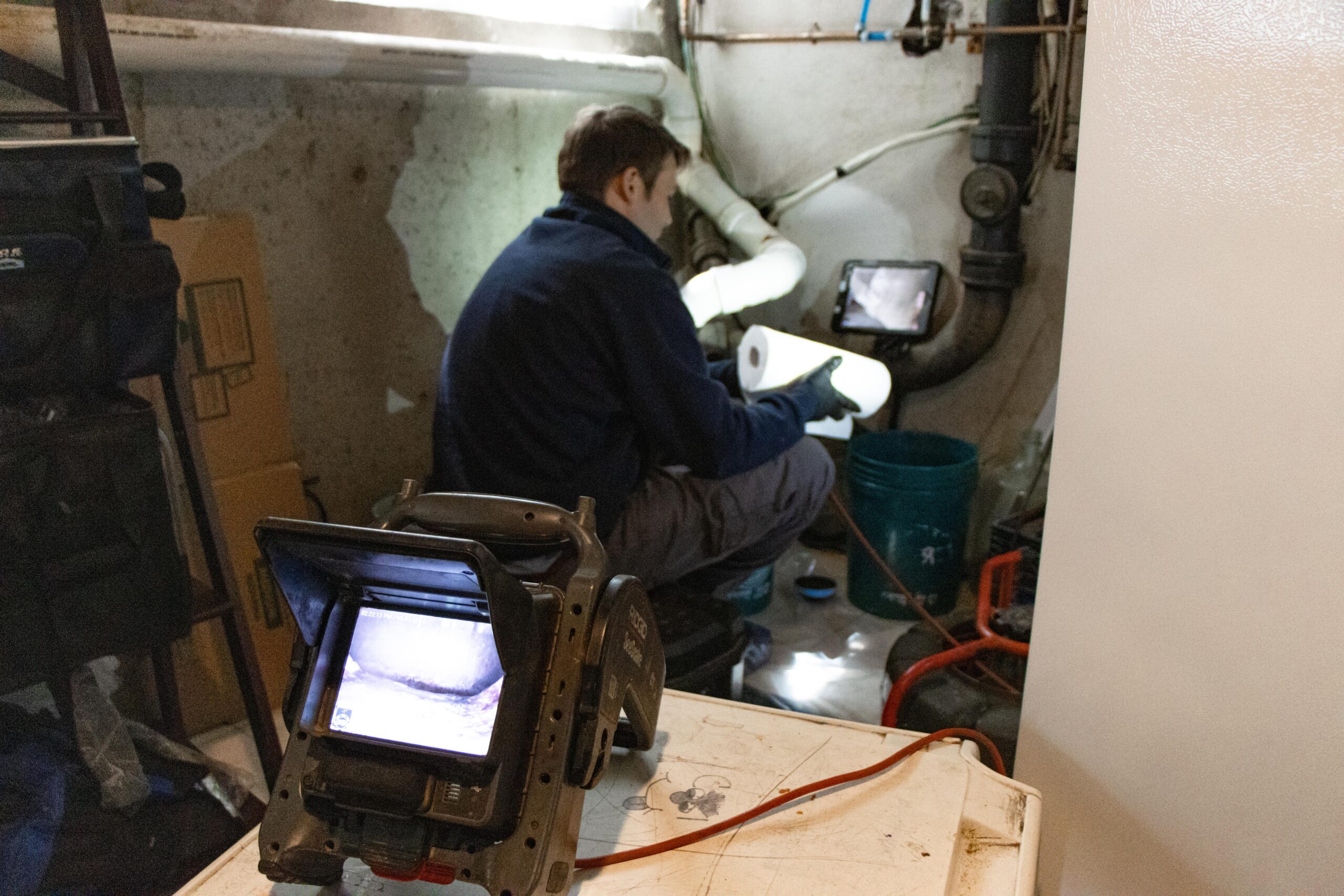 An expert plumber from 128 plumbing in Milton meticulously inspects plumbing pipes using a camera and monitor system in a basement-like setting, surrounded by various equipment and tools.