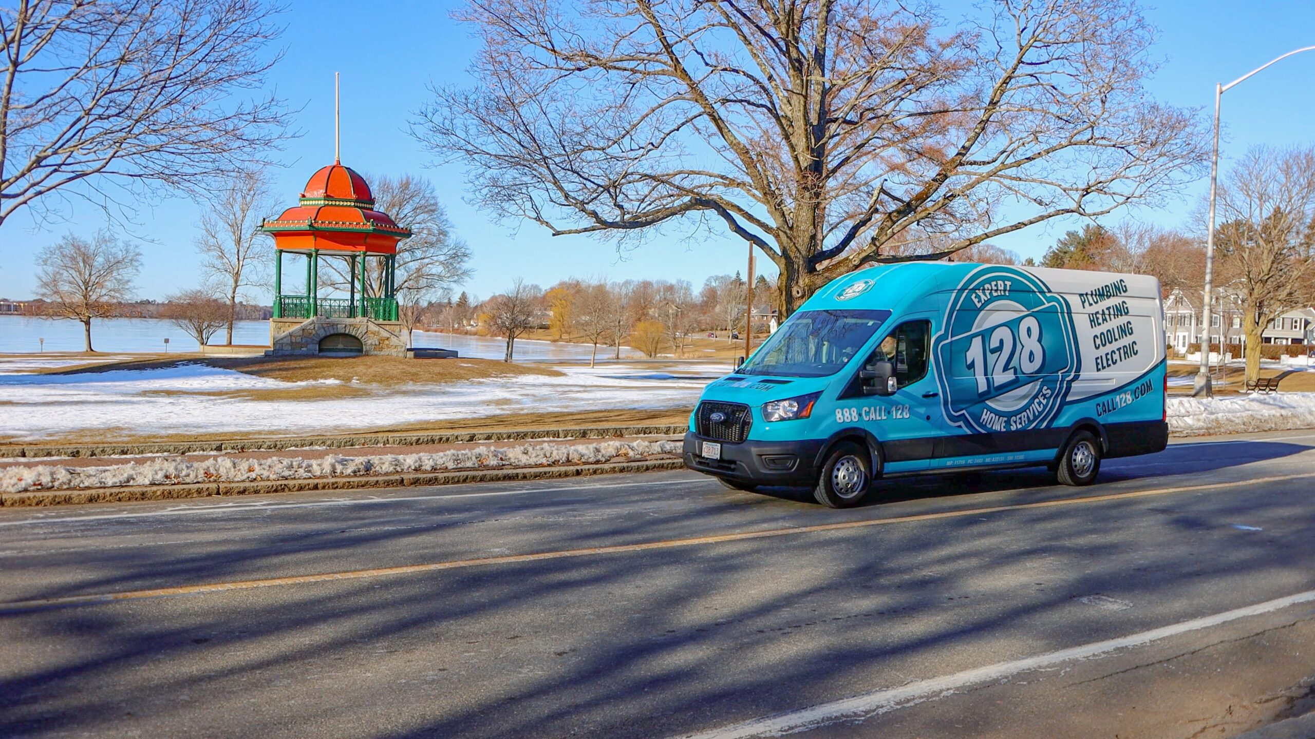 A blue and white HVAC company van from Wakefield drives on a road beside a snow-dusted park with a red-roofed gazebo and leafless trees.