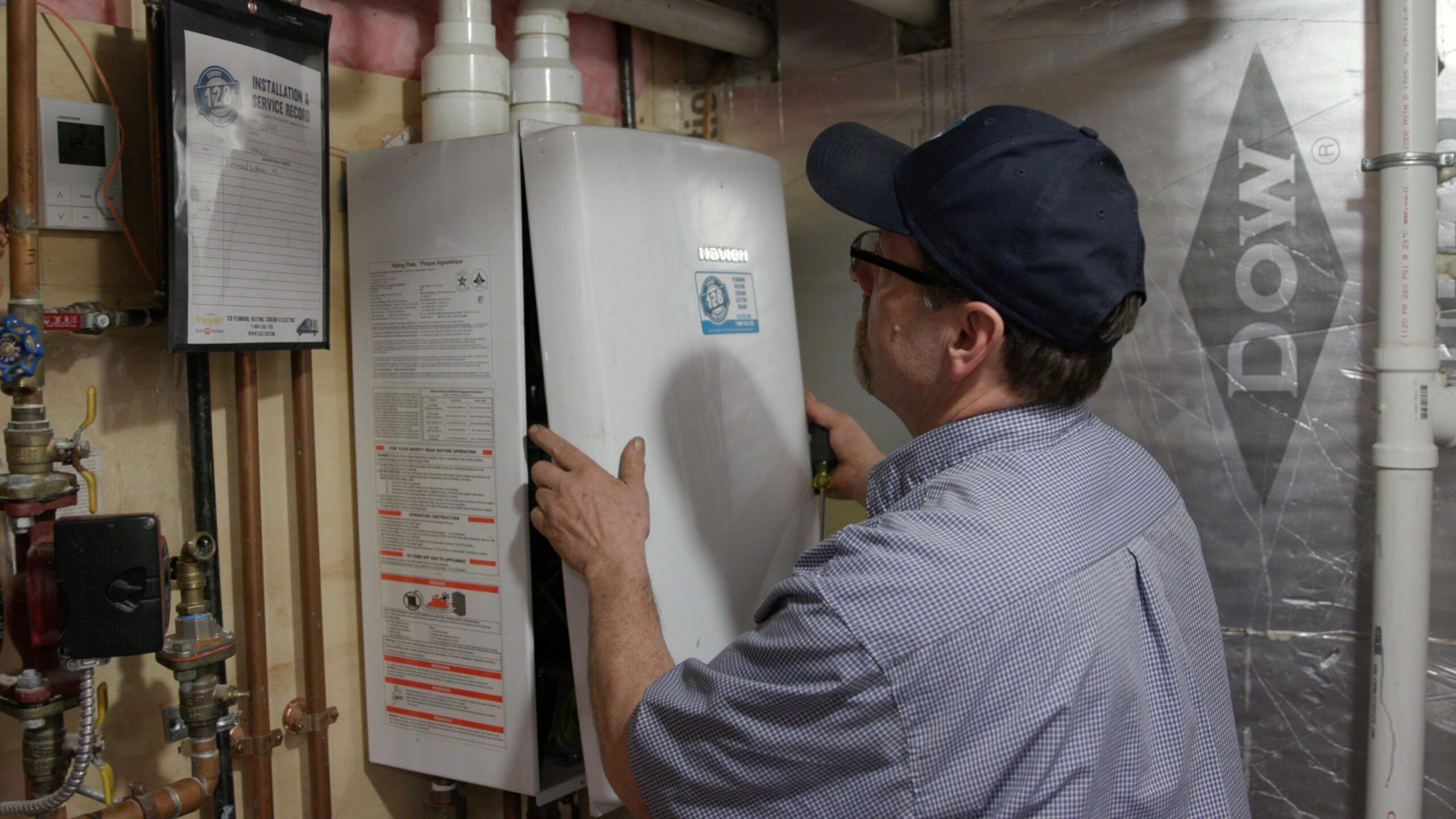 A technician in a blue cap and shirt inspects or repairs a wall-mounted tankless water heater in a utility room.
