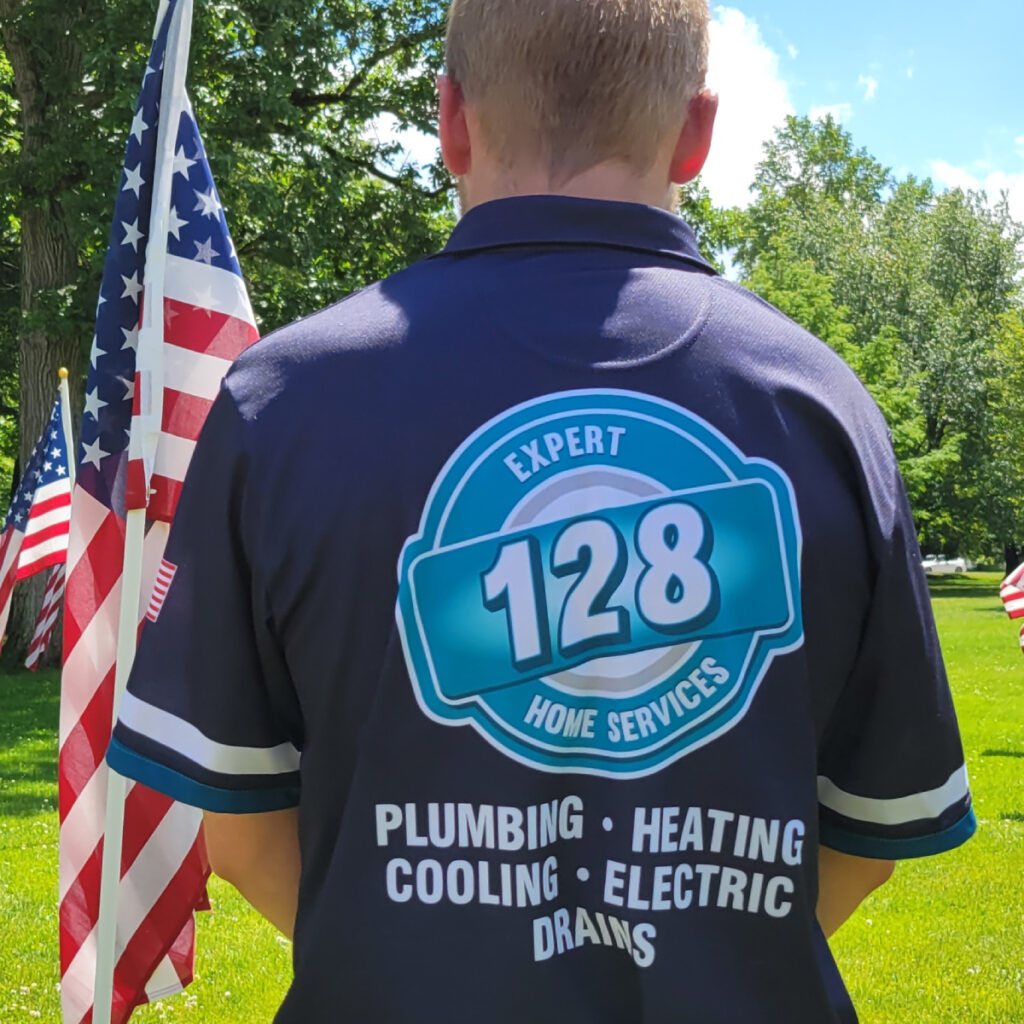 Person stands outdoors with their back to the camera, wearing a "128 Home Services" shirt listing plumbing, heating, cooling, electric, and drain work—proudly offering HVAC Services North Andover—near American flags.