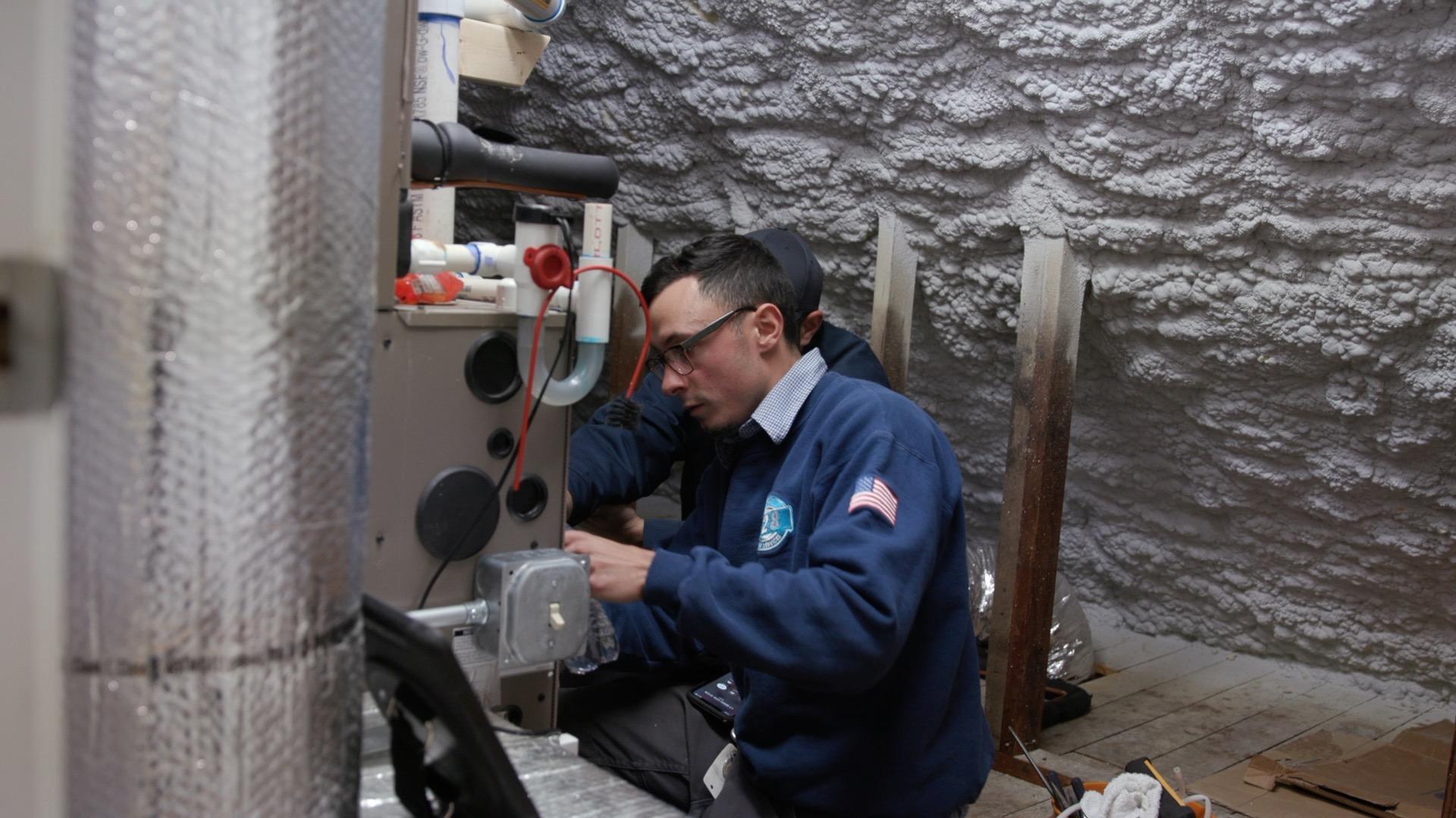A technician in a blue uniform works on HVAC equipment in an attic with spray foam insulation on the walls and ceiling.
