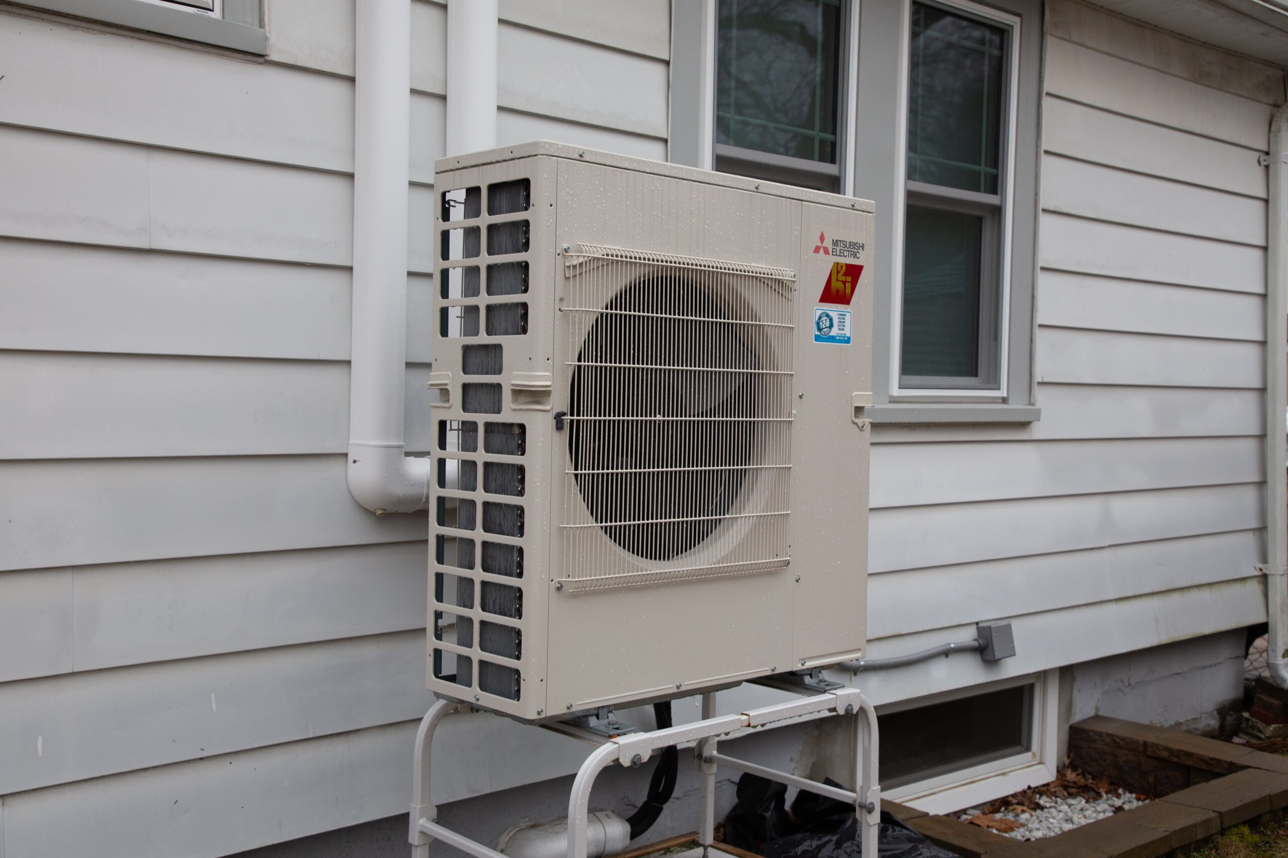 The Westford Heating outdoor air conditioning unit stands securely on a sturdy mount against the house's siding, with a window nearby.