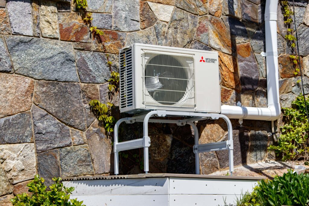 Outdoor air conditioning unit mounted on a metal stand against a stone wall, with some green plants growing nearby—a common setup where heat pumps repair may be required for optimal performance.