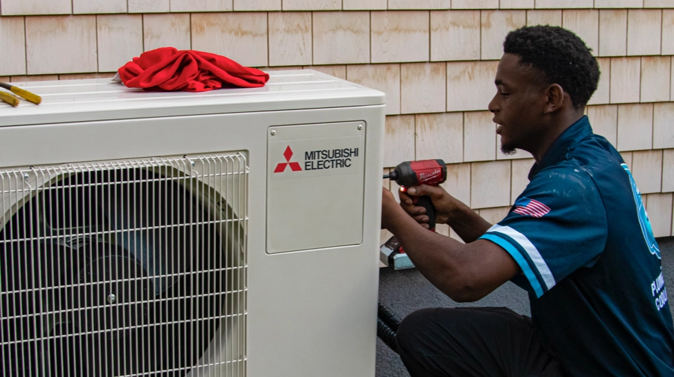 A technician uses a power drill to service a Mitsubishi Electric outdoor HVAC unit, with tools and a red cloth placed on top.